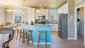 A kitchen with white cabinets, a blue island, wooden stools, a refrigerator, and a sign indicating a beach direction. - Toes In The Sand 3 Bedroom Home (Port Bolivar)
