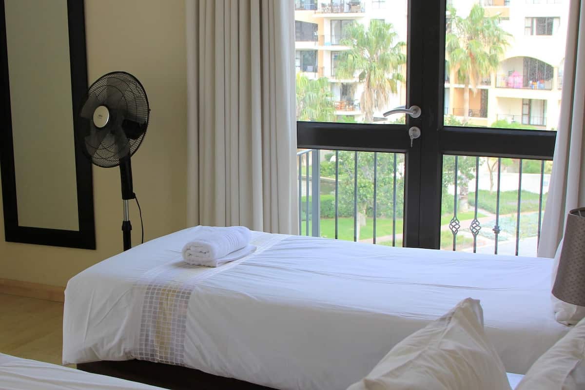 A hotel room with a bed, a fan, a window with a view of a building and palm trees.
