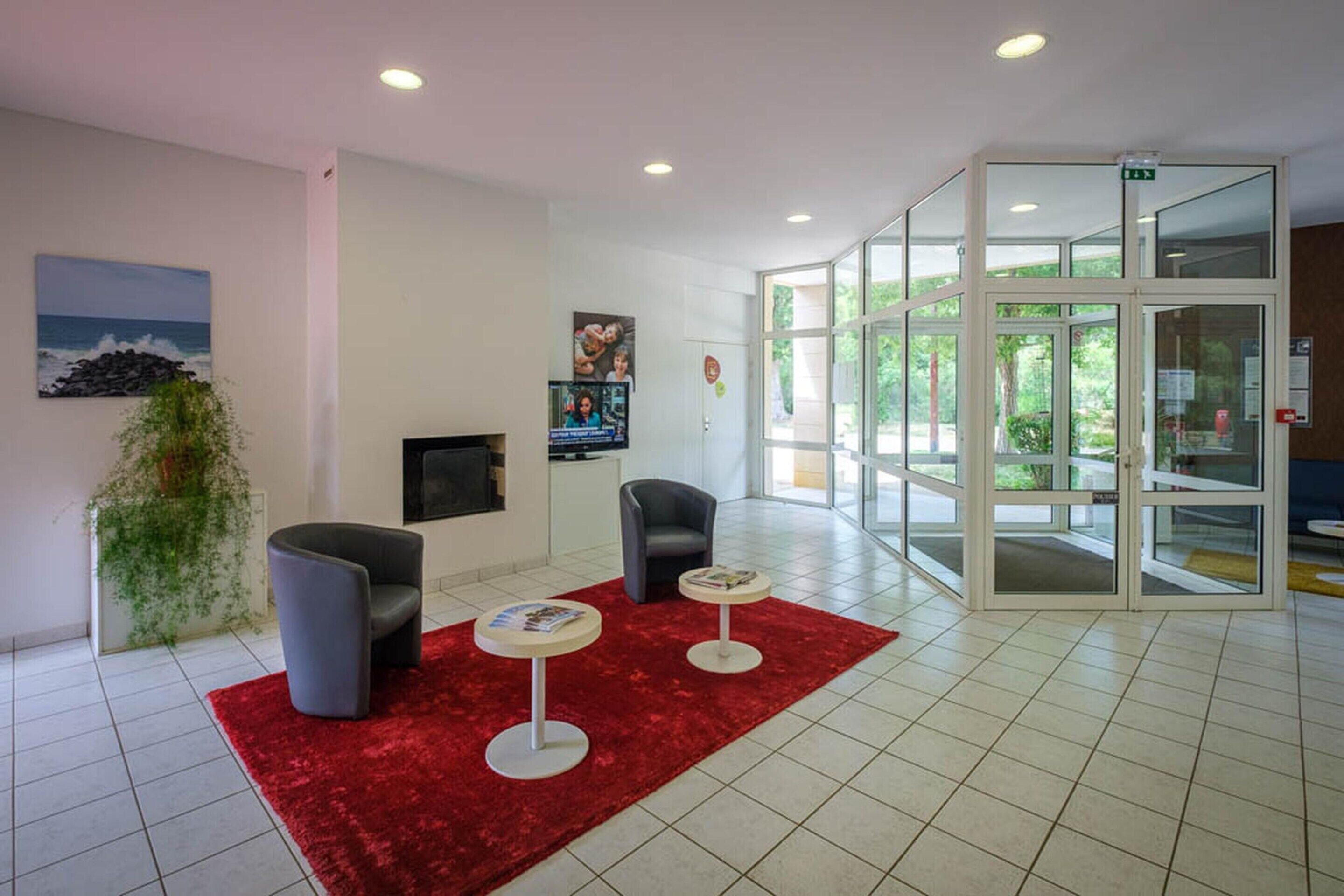 Um hall com carpete vermelho, duas poltronas, uma mesa pequena, uma televisão e uma porta de vidro que dá para o exterior.