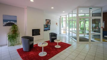 Um hall com carpete vermelho, duas poltronas, uma mesa pequena, uma televisão e uma porta de vidro que dá para o exterior.