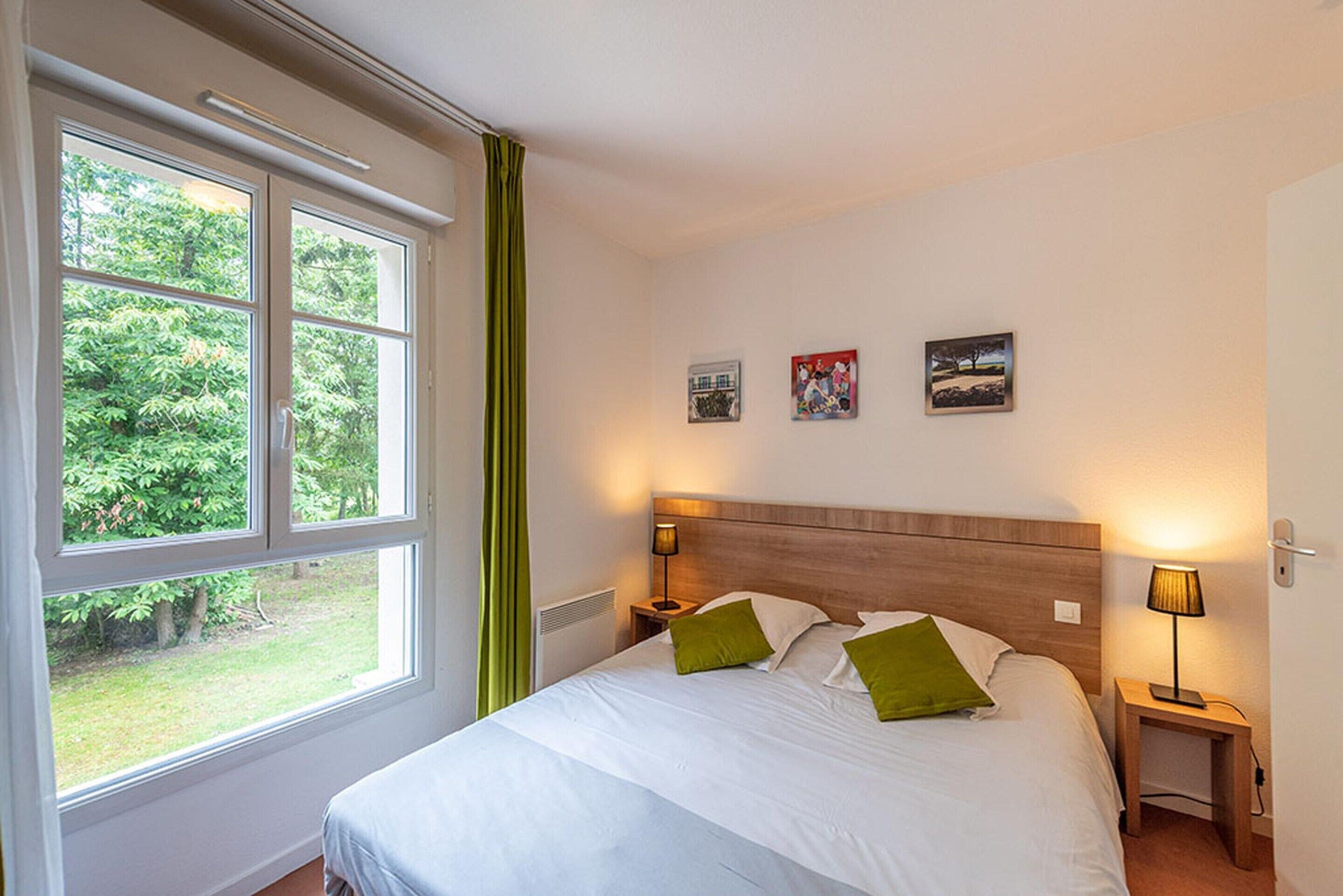 A hotel room with a large bed, two bedside tables, a nightstand, a window with curtains, and three wall-mounted pictures.