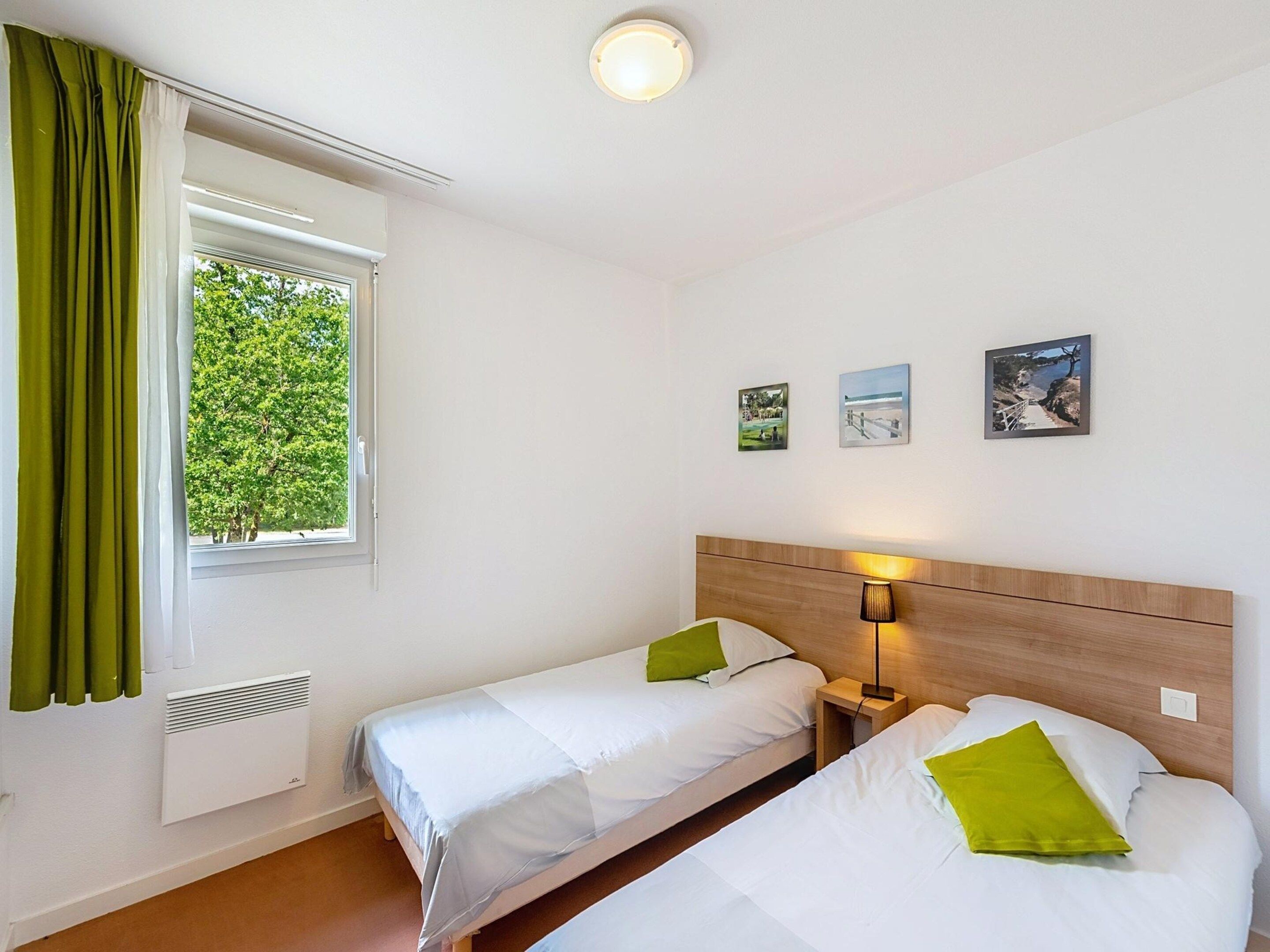 A hotel room with two beds, a window with curtains, a radiator, a nightstand with a lamp, and three framed pictures on the wall.