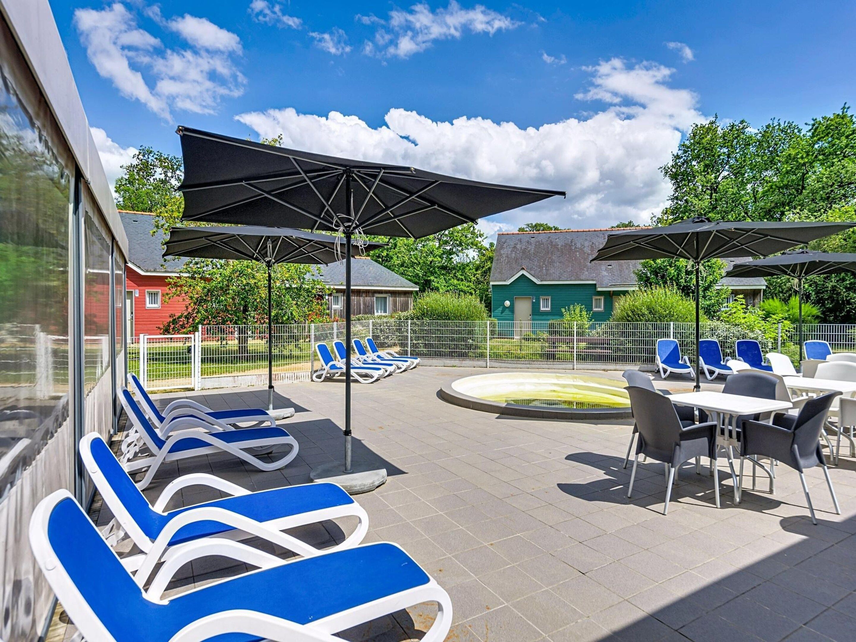 An outdoor patio area with lounge chairs and umbrellas, a pool, and a view of houses in the background.