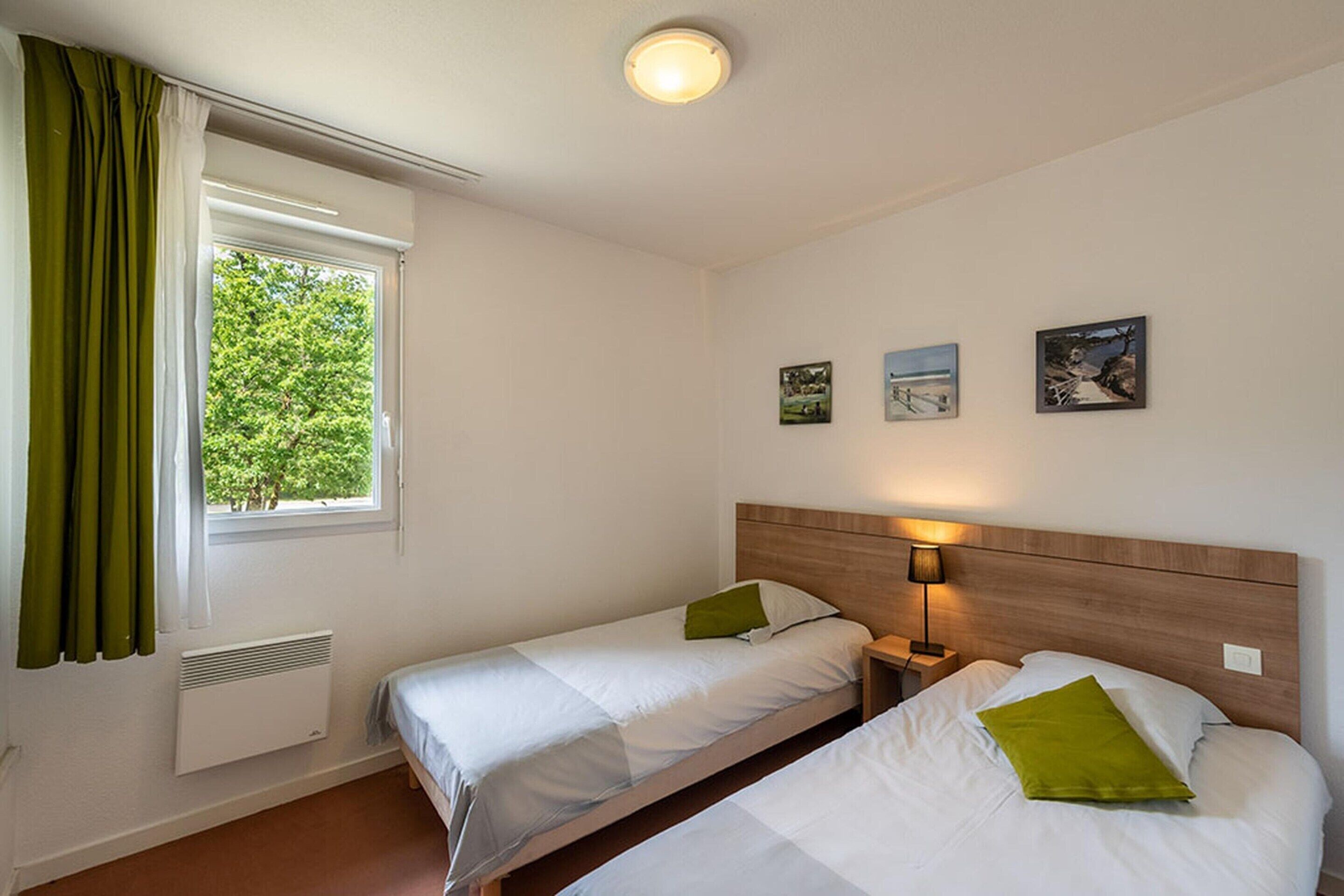 A hotel room with two single beds, a window with green curtains, a wall-mounted heater, and three framed pictures.