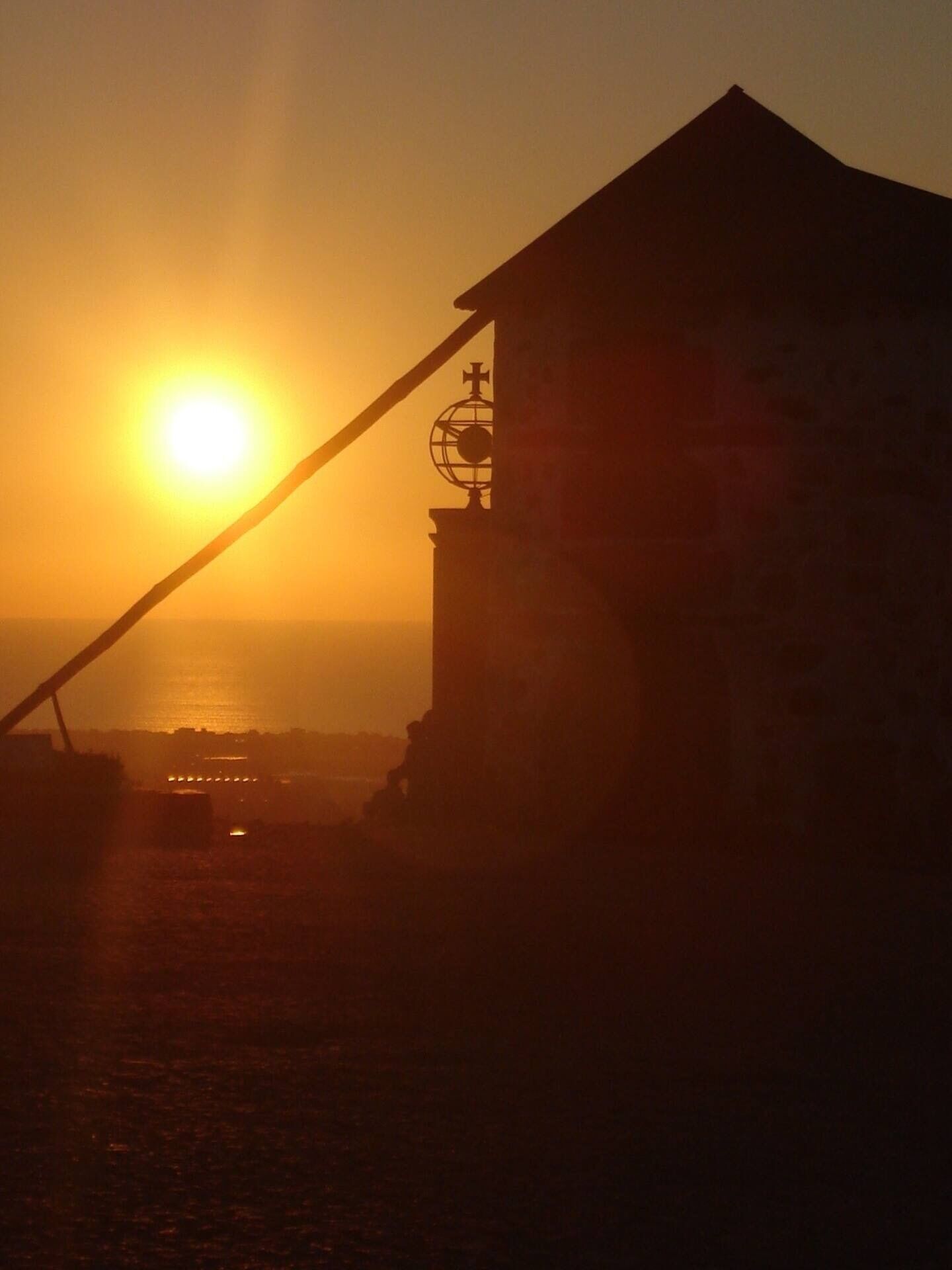 A building with a cross on top, a flagpole, and a view of the sunset over water.