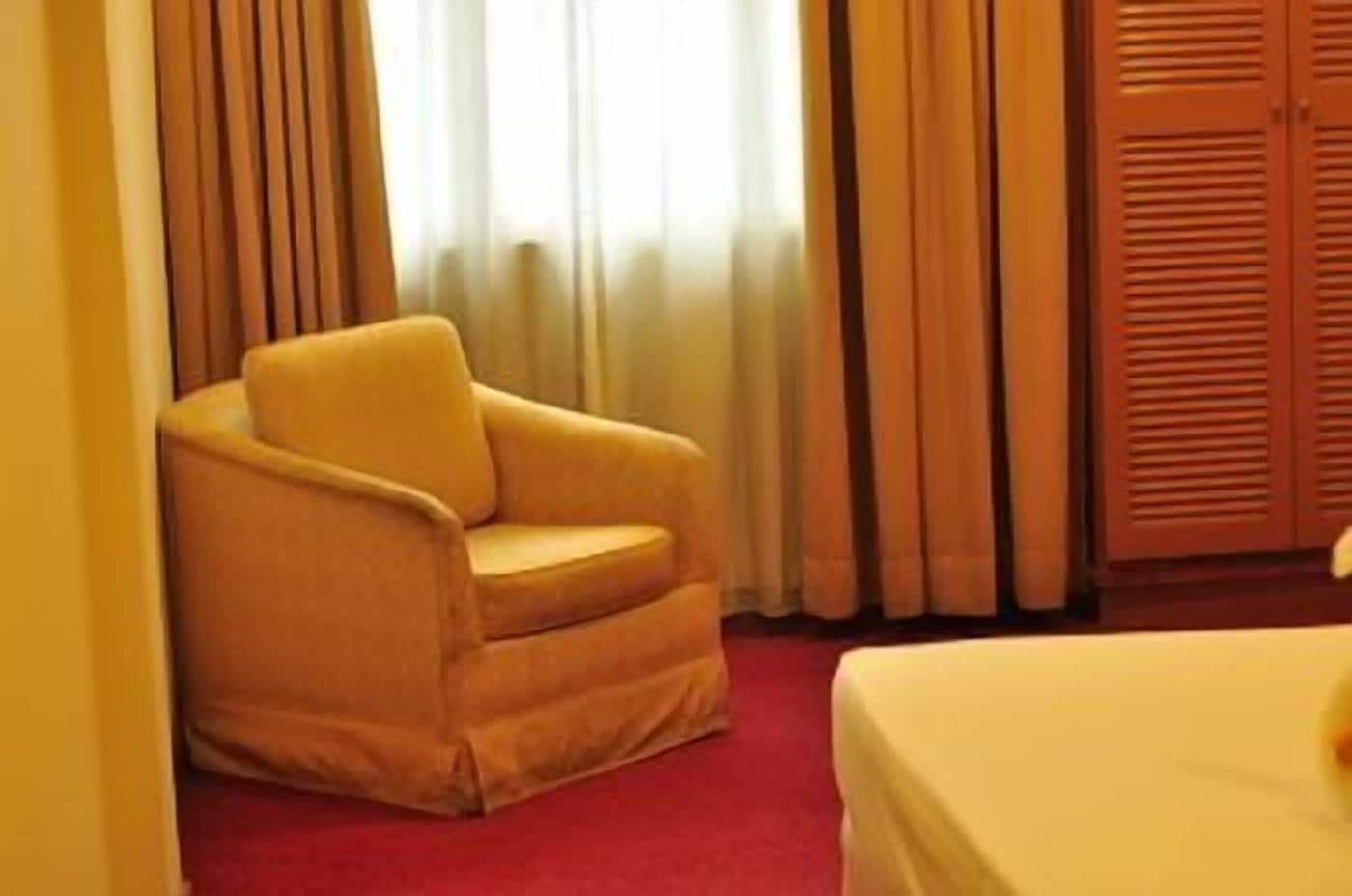 Hotellihuone, jossa on punainen matto, keltainen nojatuoli, sänky ja puinen vaatekaappi.