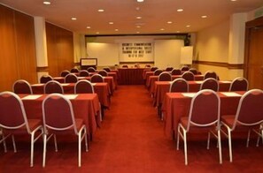 Un salón de conferencias con filas de sillas y mesas, piso alfombrado y un cartel en la pared.