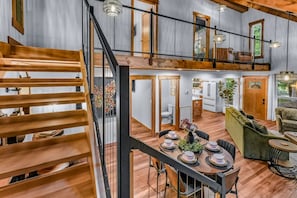 A spacious loft-style interior with a wooden staircase, a dining area with a table set for four, and a cozy seating area with a sofa and armchairs.