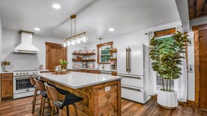 A modern kitchen with a central island, wooden bar stools, a large potted plant, and a view of the outdoors. - Dolly's Harmony Haven 3 Bedroom Cabin (Sevierville)