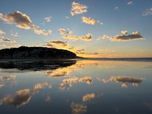Una playa tranquila con cielo despejado y nubes, un mar sereno y un acantilado rocoso al fondo.