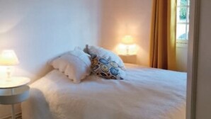 Een bed met witte lakens en opvallende kussens, een bedlampje en een raam met gordijnen.