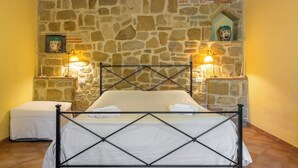 A bedroom with a stone wall, a bed with a metal frame, two bedside lamps, and a small shelf with a decorative item. - Cosy apartment in villa with A/C, WIFI, pool, TV, patio, pets allowed and panoramic view (Perugia)