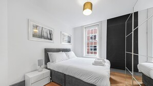 Un dormitorio moderno con cama, mesitas de noche, lámpara, cuadros enmarcados, ventana y espejo.