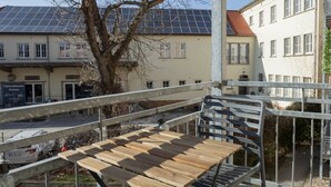 Ein Balkon mit einem Holztisch und Metallstühlen, der auf eine Straße mit Gebäuden und einem Baum blickt.