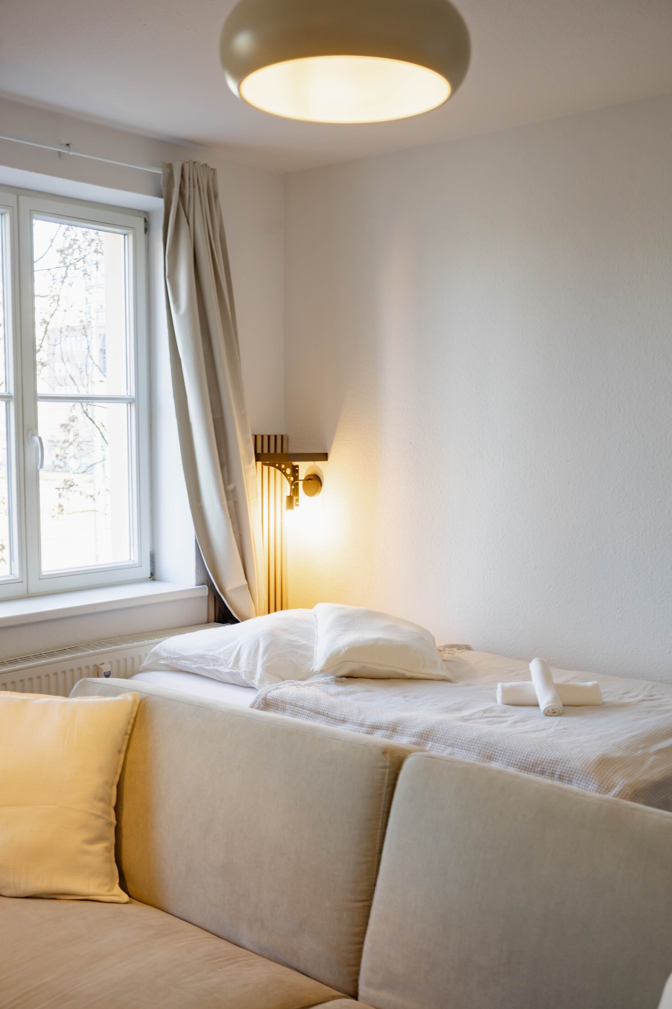 Une chambre à coucher comprenant un lit, un canapé, une fenêtre avec des rideaux et un luminaire fixé au mur.