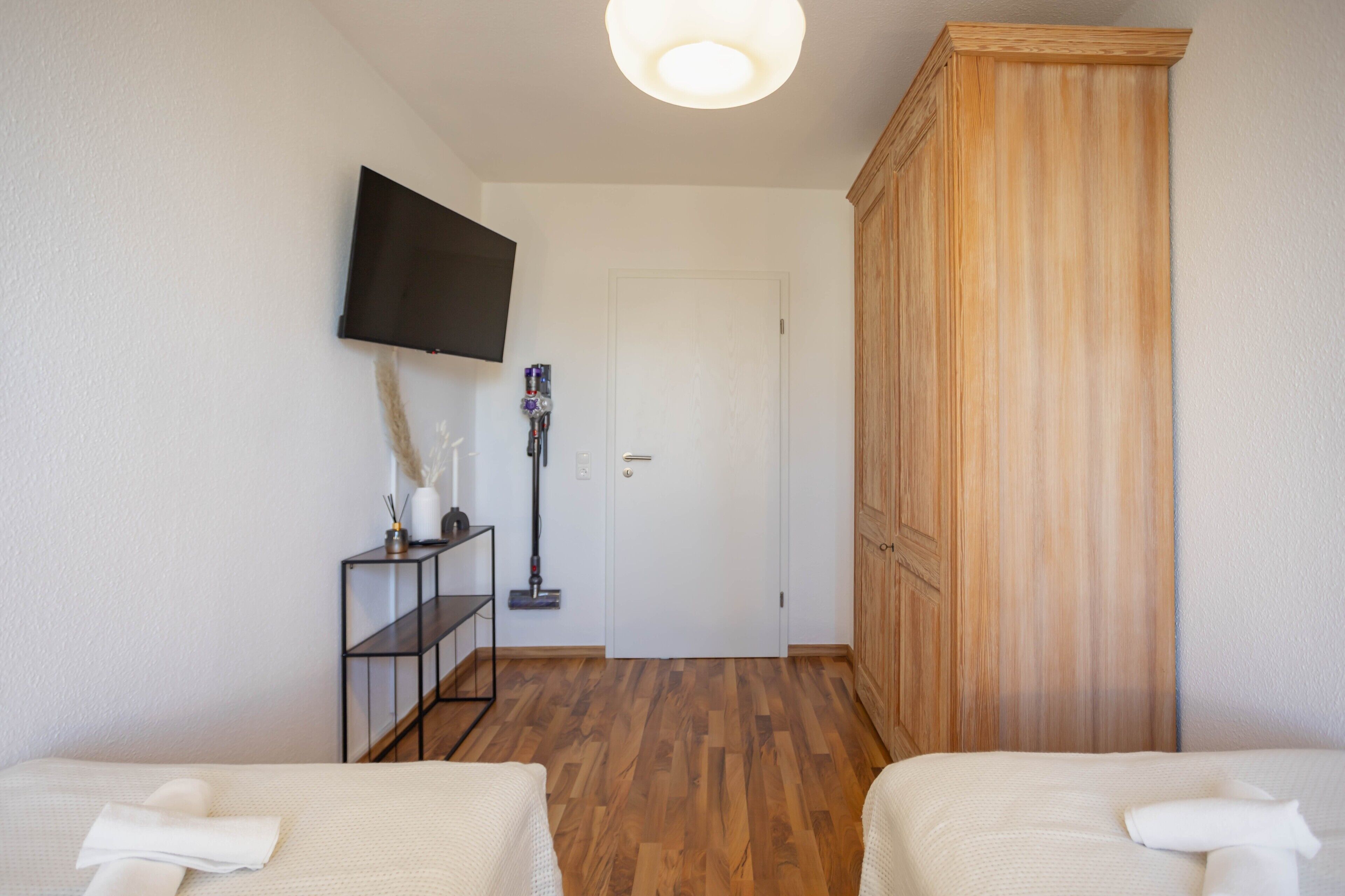 Une chambre d’hôtel avec une armoire en bois, un téléviseur fixé au mur, une étagère avec un aspirateur et deux lits.