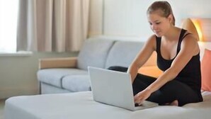 Iemand zit op een bank, met een laptop, een raam met gordijnen en een lamp op de achtergrond.