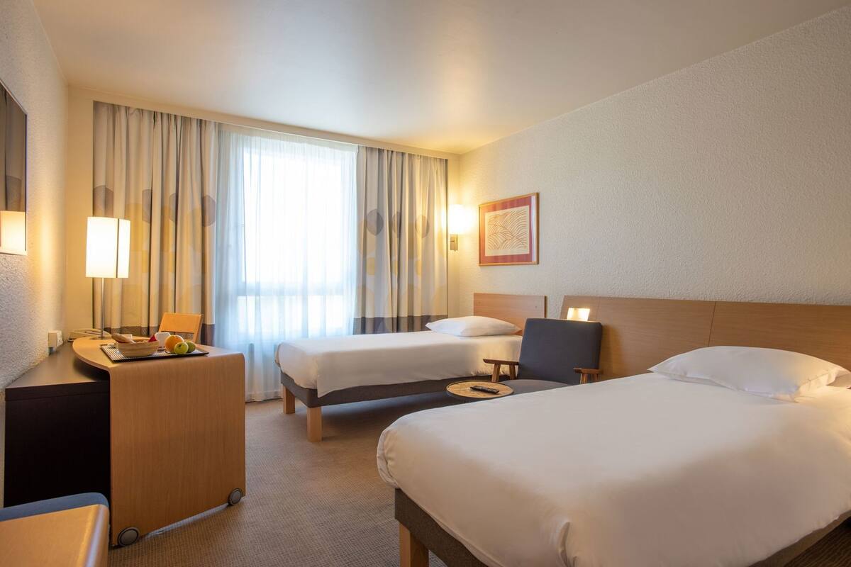 A hotel room with two beds, a desk with a lamp, a chair, and a bedside table with a fruit bowl.
