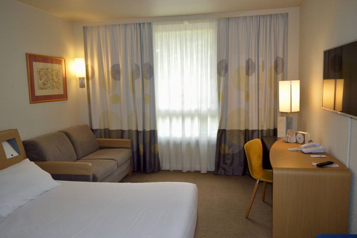 A hotel room with a bed, a sofa, a desk with a chair, a TV, and a window with curtains.