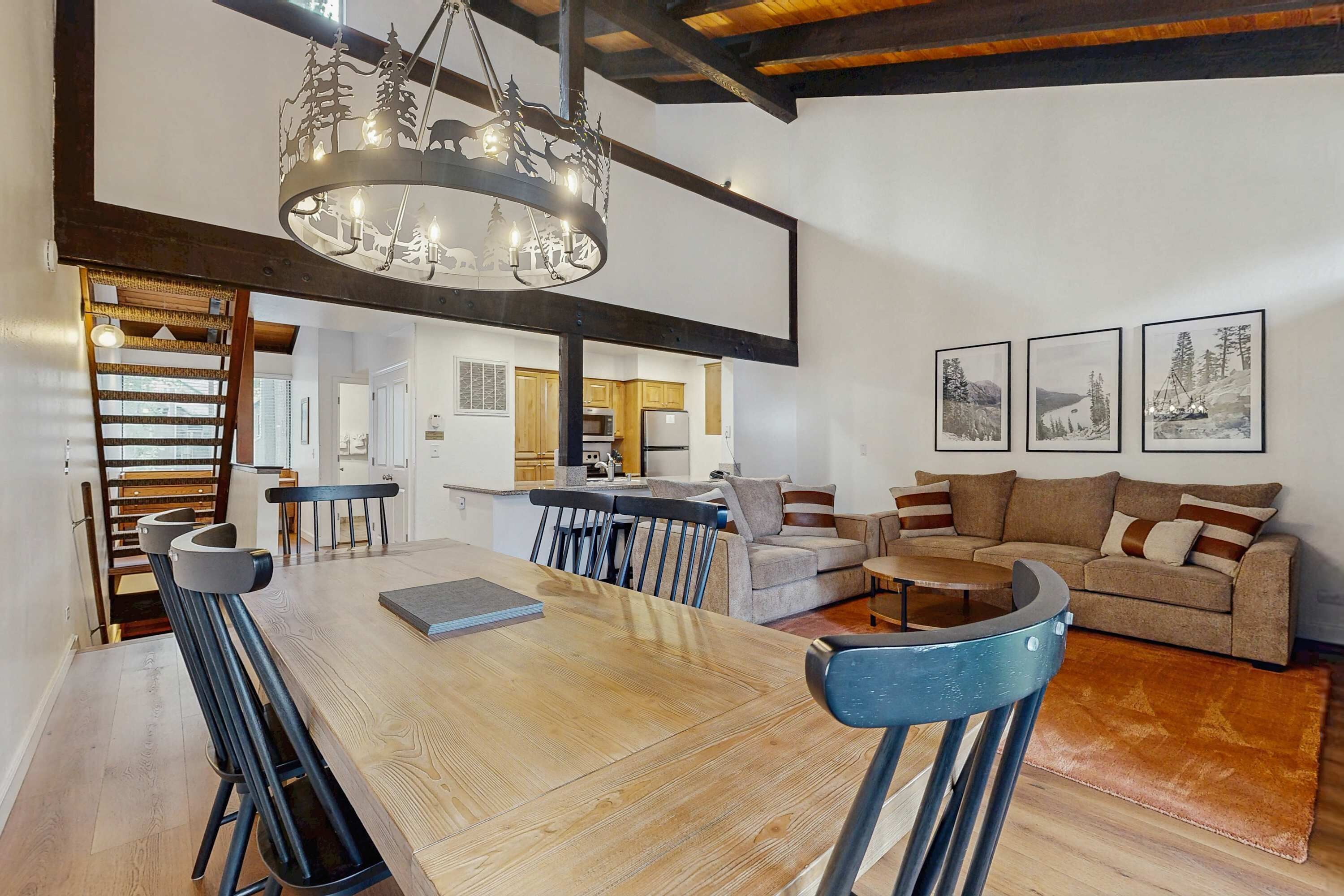 Un vaste espace dédié à la salle à manger, avec une table et des chaises en bois, une cuisine à l’arrière-plan, et un salon comprenant un canapé et des tableaux encadrés accrochés au mur.