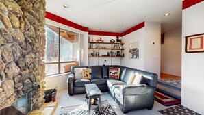 A living room with a stone wall, a sectional sofa, a coffee table, a bookshelf, and a patterned rug. - Comfortable, chic condo with mountain views - great Olympic Valley location (Squaw Valley)