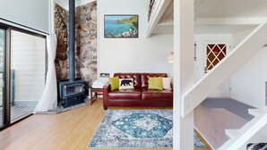 A living room with a stone wall, a fireplace, a red leather sofa, a patterned rug, and a painting of a coastal scene.