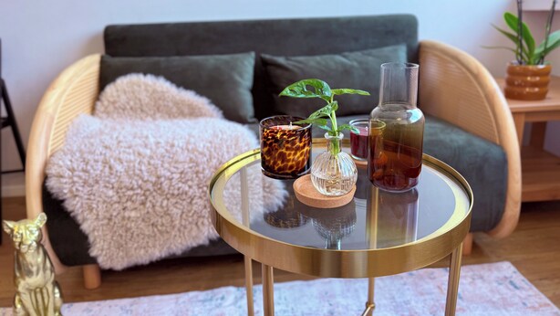 Una mesa redonda de cristal con marco dorado, un florero de cristal con una planta, una vela y una botella de líquido.