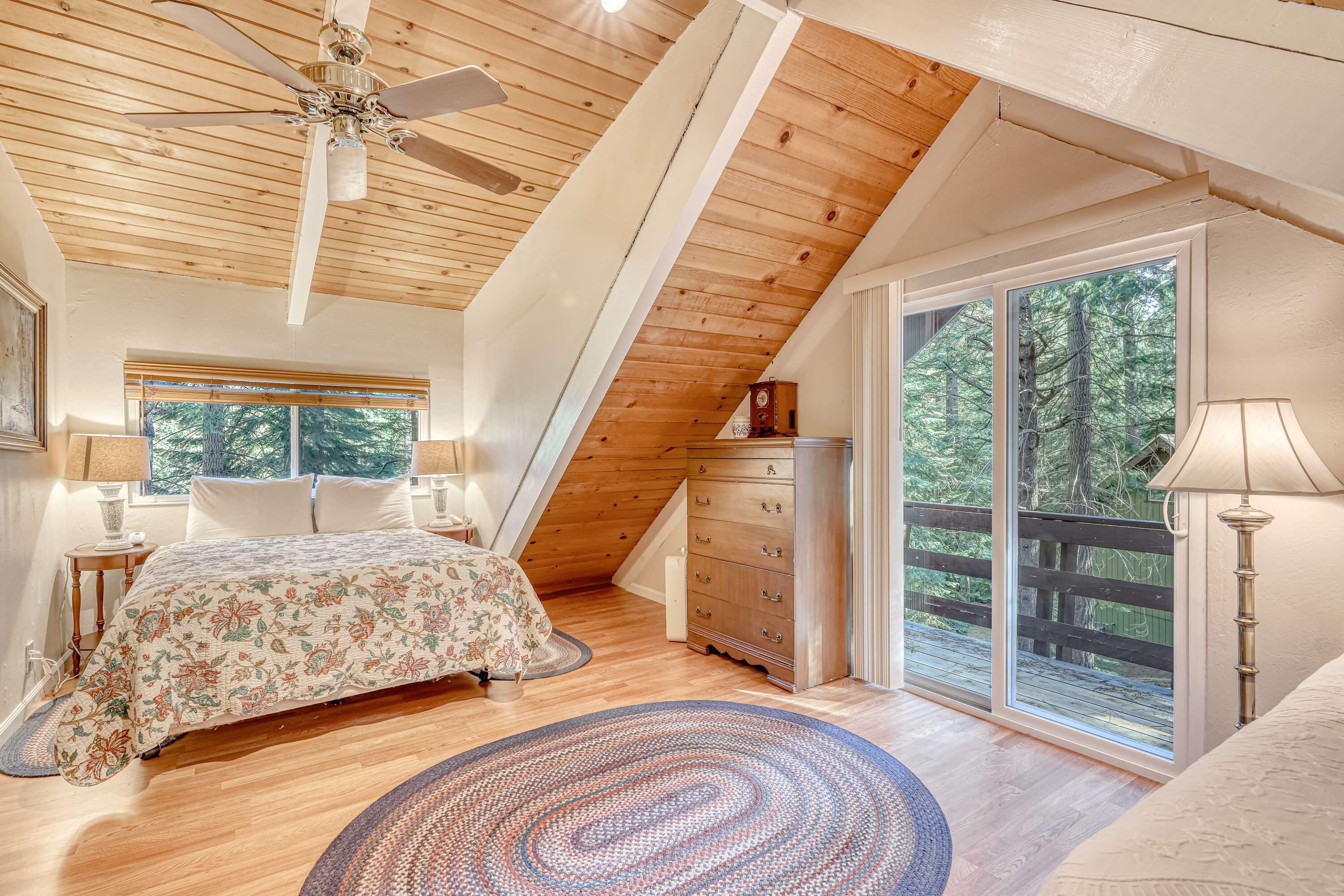 Quarto com teto de madeira, cama, penteadeira e uma janela grande com vista para árvores.