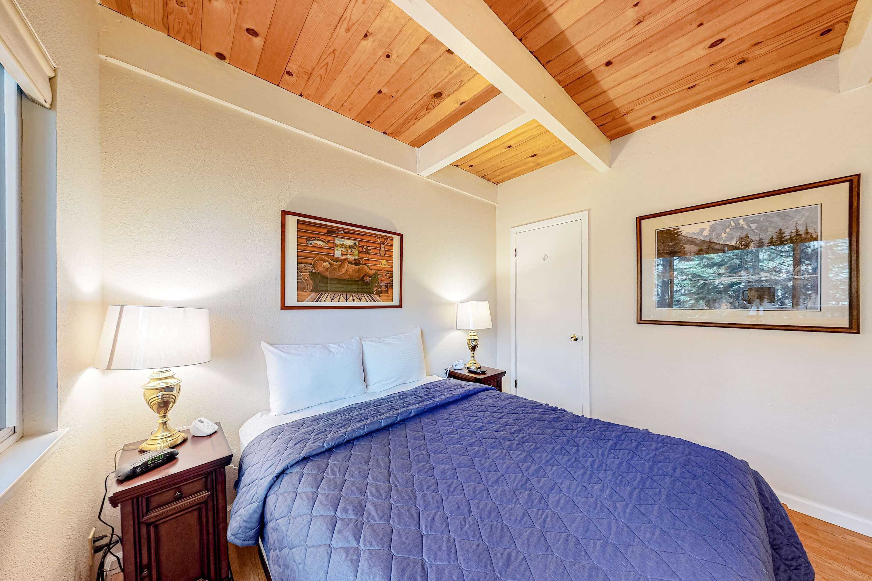 Un dormitorio con techo de madera, una cama con colcha azul, dos lámparas de noche, una mesita de noche con un control remoto y una puerta.