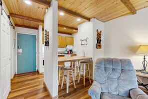 A room with a wooden ceiling, a blue door, a kitchen area with wooden cabinets, a bar stool, and a cushioned armchair. - Modern condo on the lake with pool, hot tub & balcony - short walk to the beach (South Lake Tahoe)