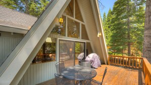 A wooden deck with a table and chairs, a covered grill, and a glass door leading to an interior space. - A-frame home with large deck, game room, wood fireplace & near the lake - dogs ok (Carnelian Bay)