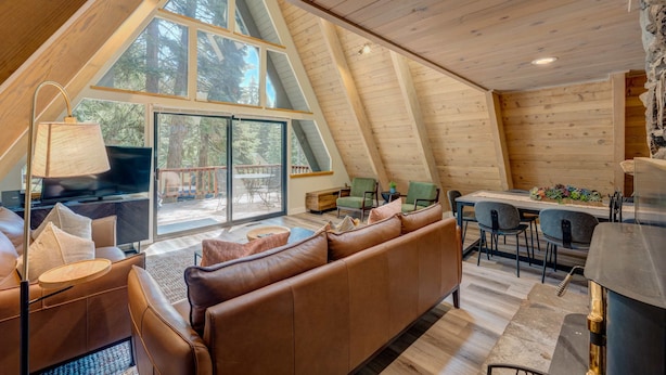A cozy, modern loft-style room with a large window, a sofa, a dining table, and a kitchen area. - A-frame home with large deck, game room, wood fireplace & near the lake - dogs ok (Carnelian Bay)