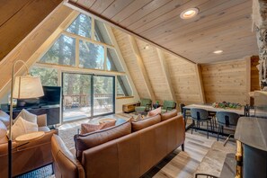 A cozy, modern loft-style room with a large window, a sofa, a dining table, and a kitchen area. - A-frame home with large deck, game room, wood fireplace & near the lake - dogs ok (Carnelian Bay)