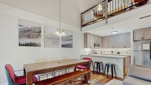 Ein Essbereich mit Holztisch, roten Stühlen und einer Bank. Zwei Gemälde hängen an der Wand, eine Küche mit Kühlschrank, Mikrowelle und Spüle sowie eine Treppe, die in einen oberen Bereich führt.