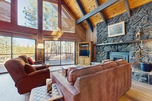 A spacious living room with a stone fireplace, wooden beams, and large windows overlooking a deck. - Beautiful, spacious mountain getaway close to skiing & hiking (South Lake Tahoe)