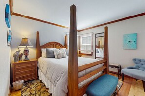 Ein Schlafzimmer mit einem Himmelbett, einem Holzkopfstück, Nachttischen, einer Lampe, einem Stuhl und einem Fenster mit Jalousien.