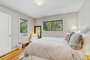 Ein Schlafzimmer mit einem großen Bett, zwei Fenstern, einem Nachttisch, einem Sessel und einer Lampe.
