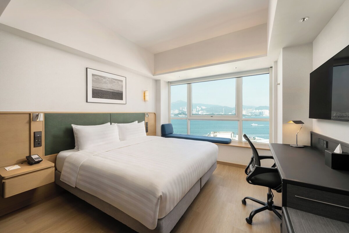 Ein modernes Hotelzimmer mit einem großen Bett, einem Schreibtisch, einem Sessel, einem Fernseher und einem Fenster mit Blick auf die Stadt.