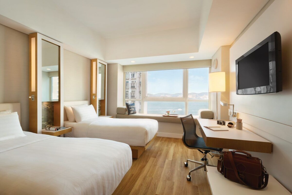 Ein Hotelzimmer mit zwei Betten, einem Schreibtisch, einem Sessel, einem Fernseher und einem Fenster mit Blick.