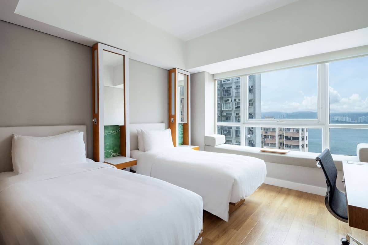 A hotel room with two beds, a desk, a chair, a large window with a city view, and a mirror.