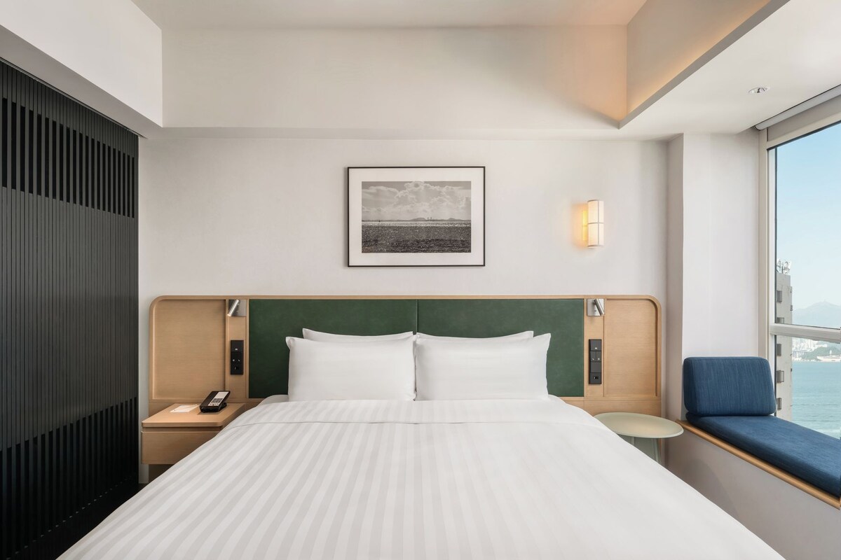 Ein Hotelzimmer mit einem großen Bett, einem Nachttisch, einer Fensterbank, einer Wandlampe, einem Fenster mit Aussicht und einem gerahmten Bild über dem Bett.