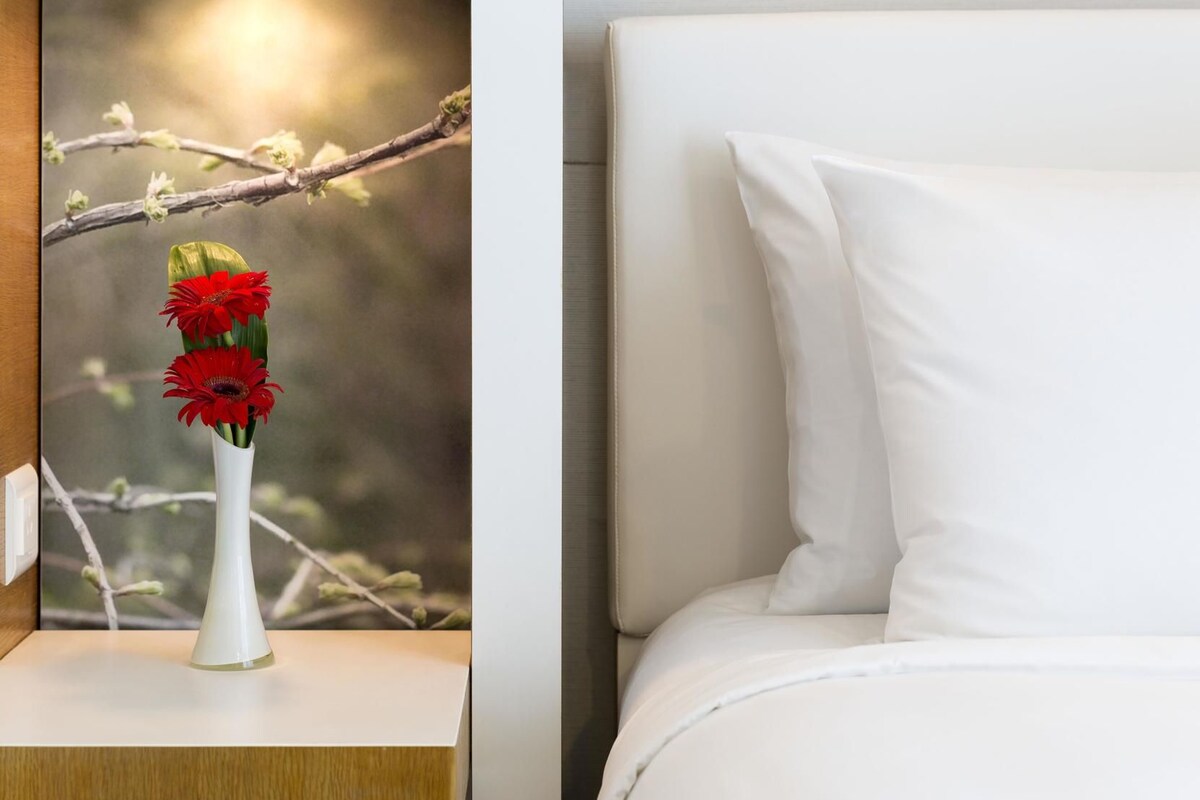 Eine weiße Vase mit roten Blumen auf einem Nachttisch neben einem Bett mit weißer Bettwäsche.