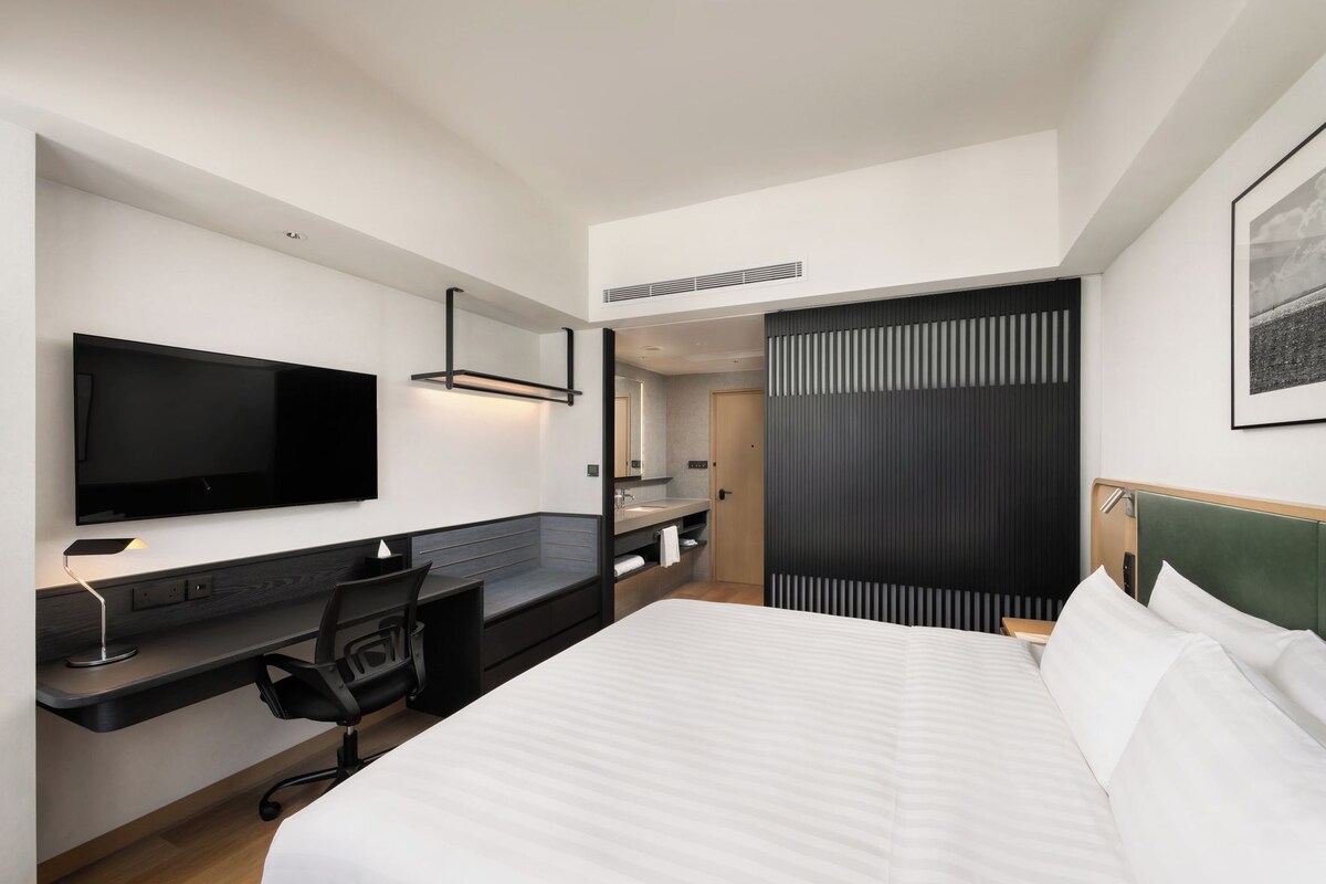 Ein modernes Hotelzimmer mit Flachbildfernseher, Schreibtisch mit Stuhl, Bett mit weißen Bettwäsche und großem Fenster.