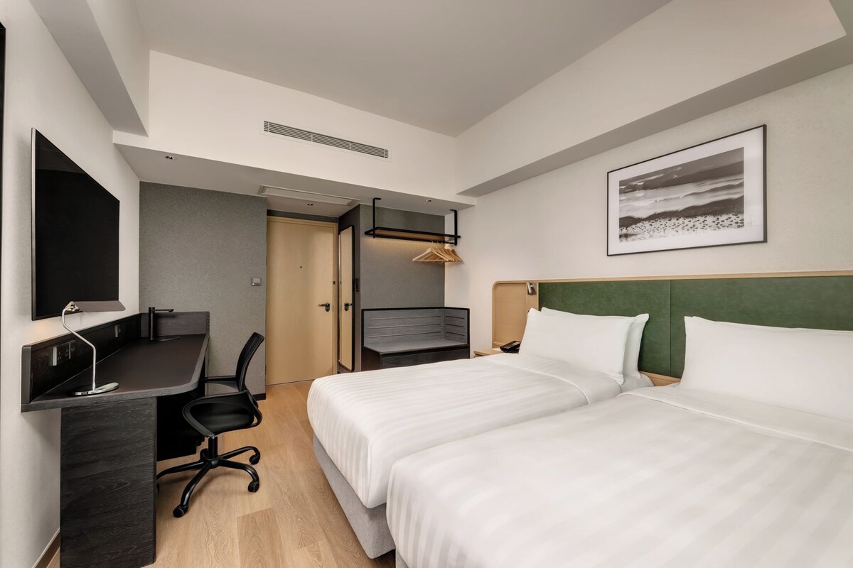 Ein Hotelzimmer mit zwei Betten, einem Schreibtisch, einem Sessel, einem Kleiderschrank und einem gerahmten Bild an der Wand.