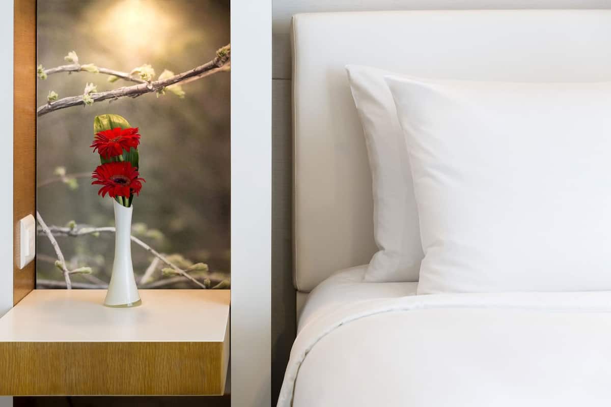 A white bedside table with a vase of red flowers and a white bed with pillows.