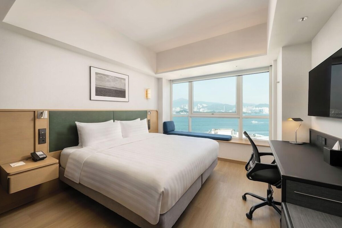 Ein Hotelzimmer mit Bett, Schreibtisch, Stuhl und einem großen Fenster mit Blick auf die Stadt.