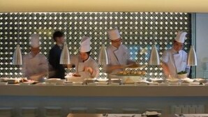 Une cuisine où des chefs en uniforme blanc s’affairent, un mur orné de motifs et un buffet disposé pour le service.