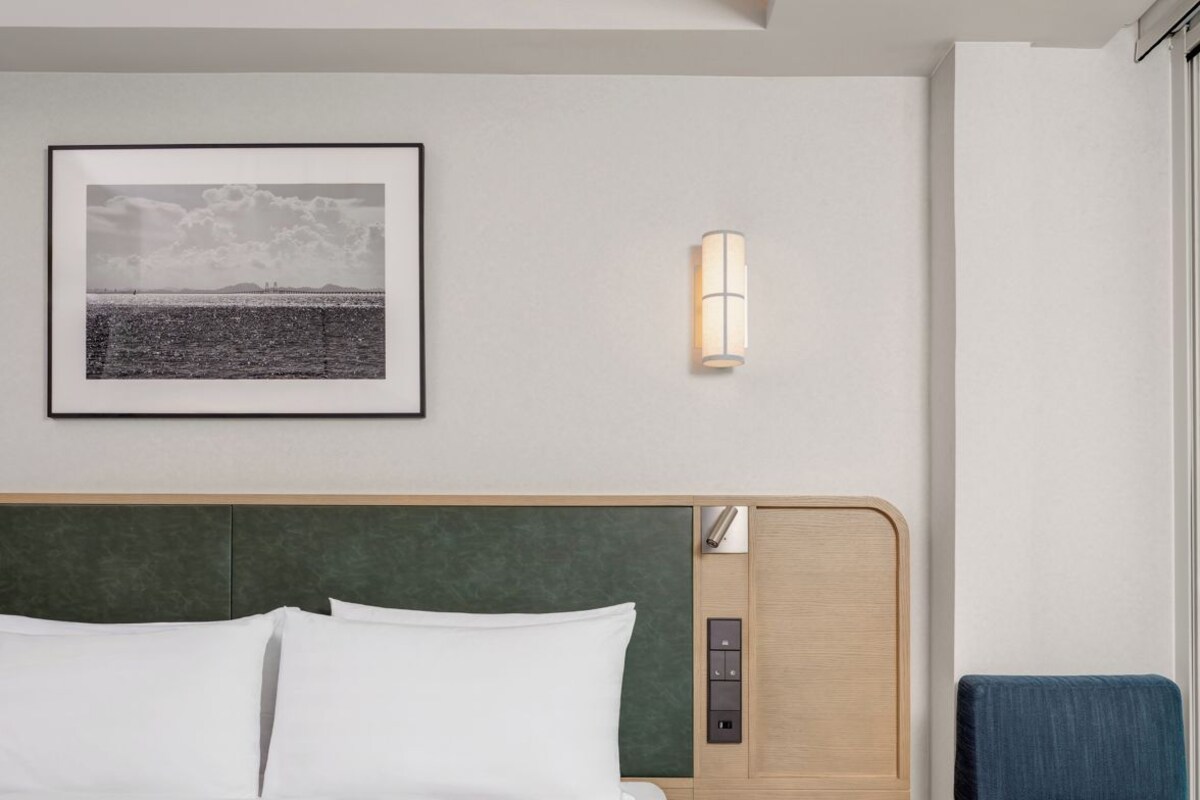 Ein Hotelzimmer mit einem Bett, einem kleinen runden Tisch, einer Sitzecke mit einem blauen Kissen, einer Wandleuchte und einem gerahmten Bild an der Wand.