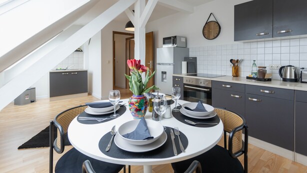 Une cuisine moderne avec des meubles de couleur sombre, un crédence carrelée de couleur blanche et un coin repas avec une table ronde prévue pour quatre personnes.