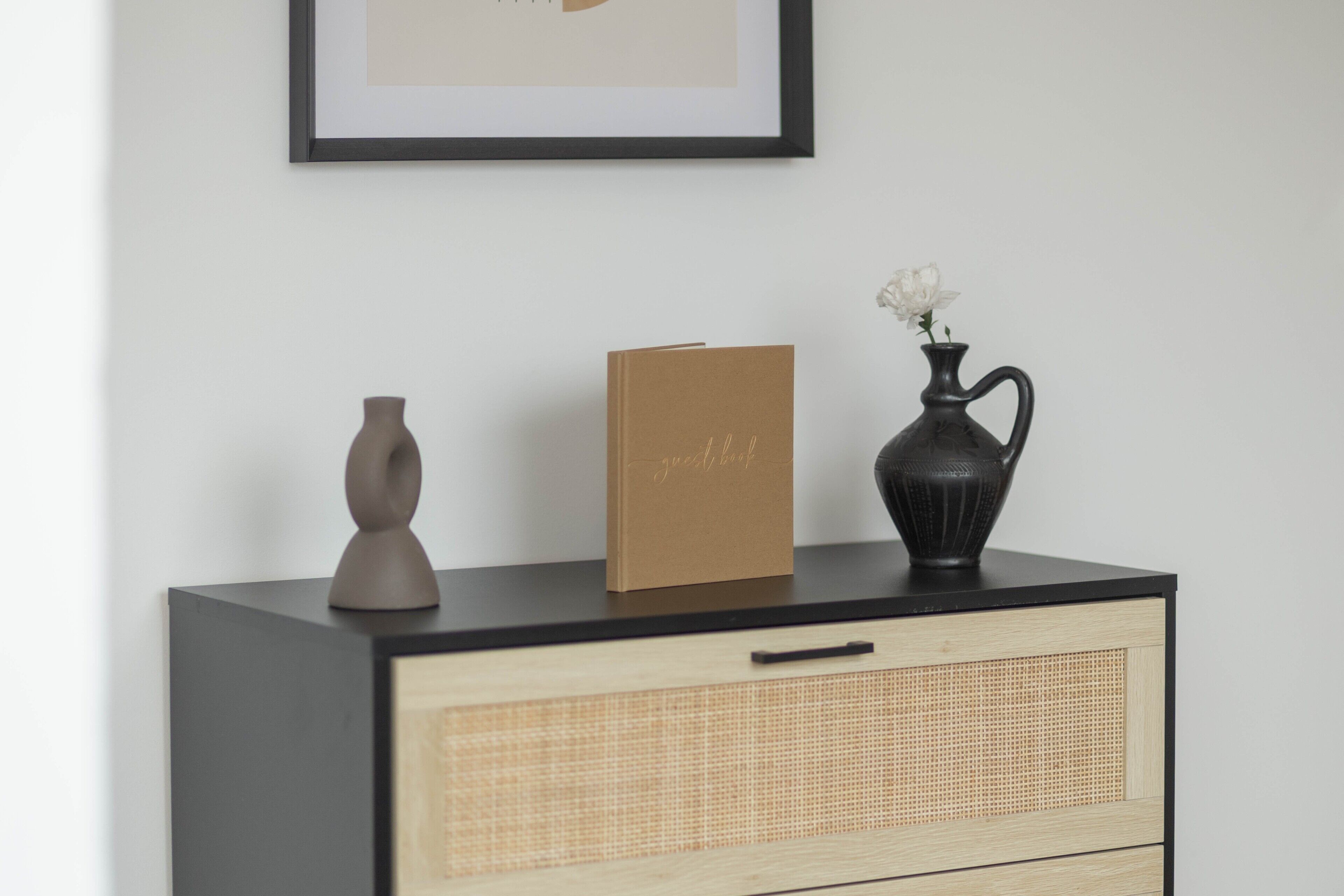 Un comò nero con pannello frontale intrecciato, un biglietto marrone, un vaso nero con fiori bianchi e un quadro appeso al muro.